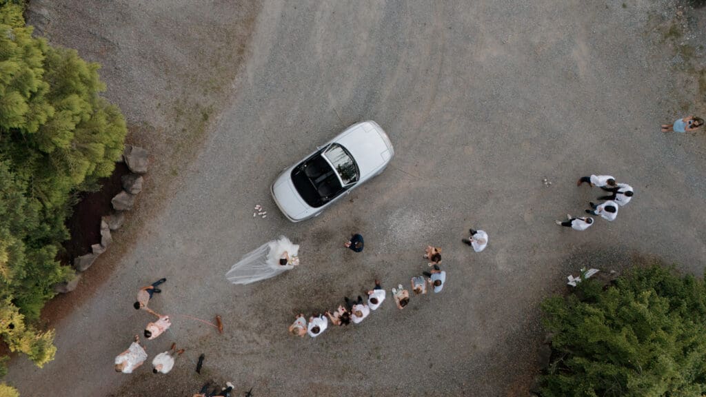 An overhead view from the drone video captured by Hayley Zumkeller photography and videography team from Victoria BC. This image captures the wedding party sending the groom and bride and their pets off following the reception in a vintage car with cans trailing behind. Taken in Nanaimo BC on Vancouver Island.