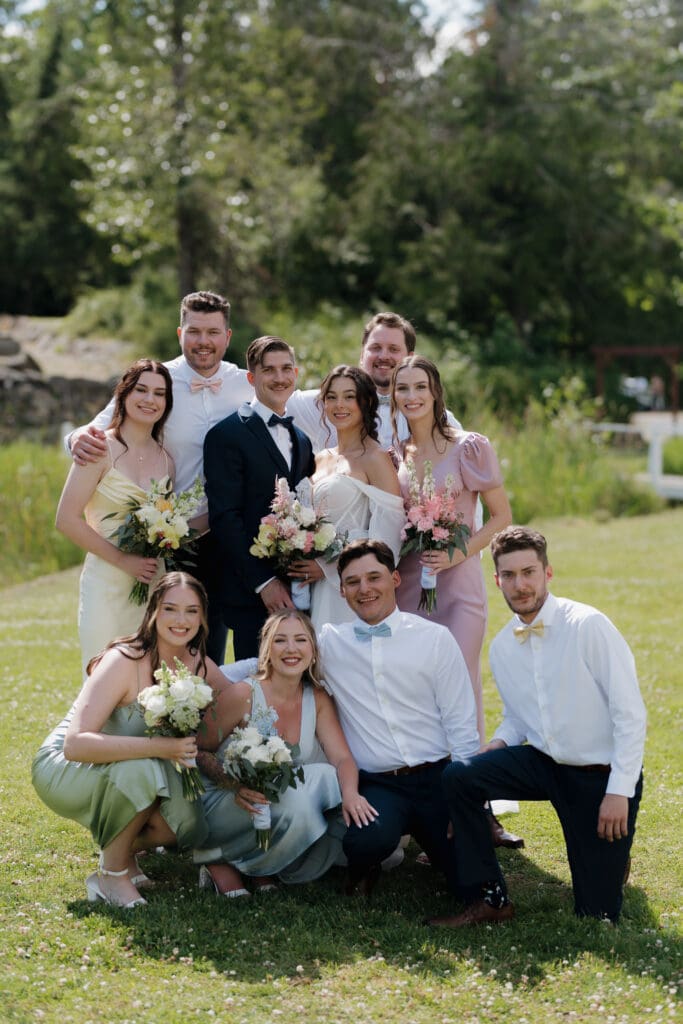 Hayley Zumkeller photography and videography team from Victoria BC capture a wedding party surrounding the groom and bride following their Nanaimo outdoor wedding ceremony. The bridesmaid's gowns are in a variety of pastel colours while the groomsman are coatless. The lush surroundings of Puska Properties show in the background.