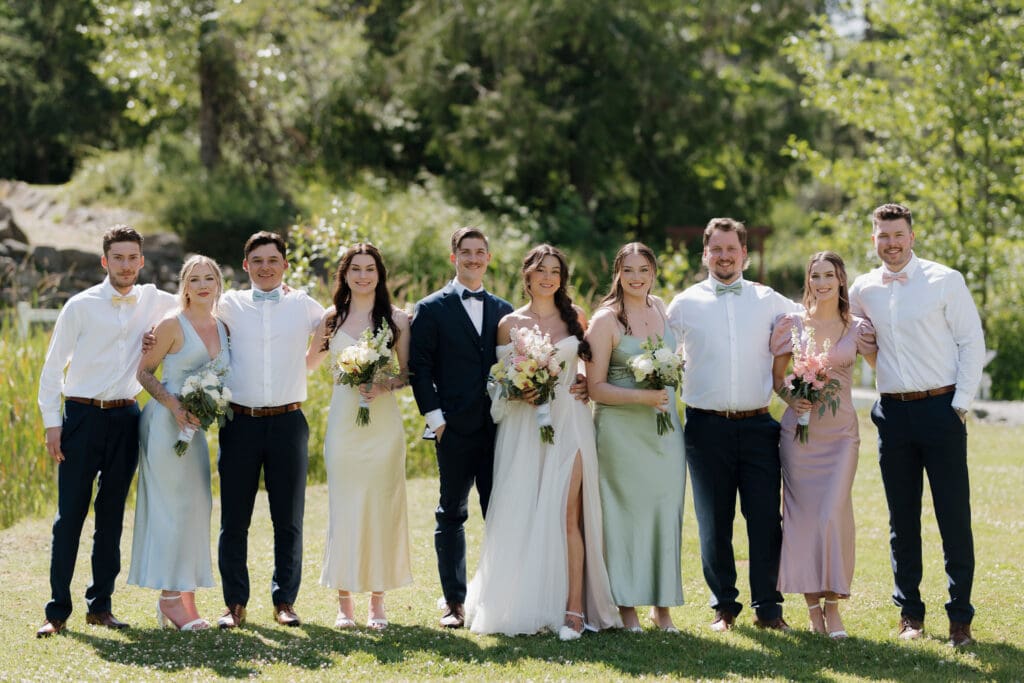 Hayley Zumkeller photography and videography team from Victoria BC capture a wedding party surrounding the groom and bride following their Nanaimo outdoor wedding ceremony. The bridesmaid's gowns are in a variety of pastel colours while the groomsman are coatless. The lush surroundings of Puska Properties show in the background.