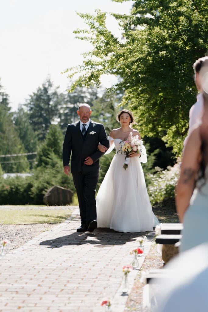 Hayley Zumkeller photography and videography team from Victoria BC capture a bride walking down the aisle in her flowing, whimsical dress supported by the father of the bride at the start of the Nanaimo outdoor wedding ceremony. The cement walkway is lined with tiny flower vases with seasonal, colourful flowers courtesy of Cora Lynn Flowers.
