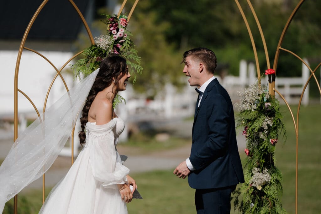 Hayley Zumkeller photography and videography team from Victoria BC capture the excitement of a newly married bride and groom at Puska Properties in Nanaimo. Both are smiling widely at each other in amazement of this epic moment. The floral arch from Cora Lynn is behind them.