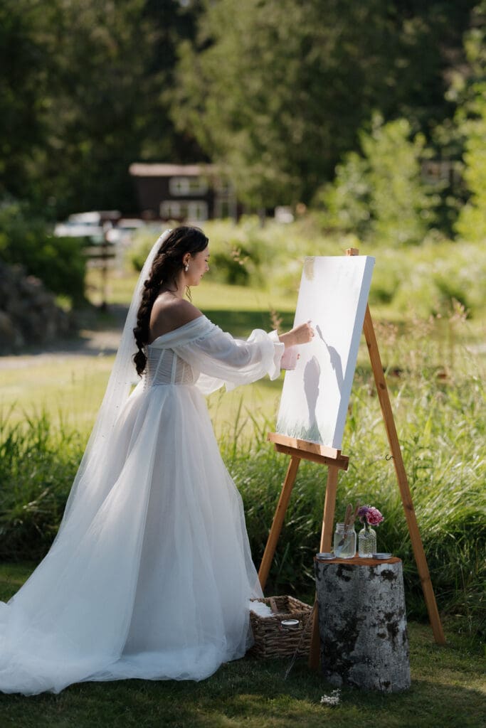 Hayley Zumkeller photography and videography team from Victoria BC capture a bride among the lush greenery at Puska Properties in Nanaimo BC. She is drawing on an easel.