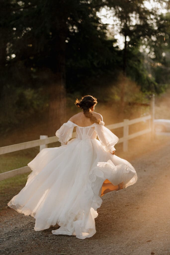 A bride surrounded by golden hour light runs after the vintage wedding car starts to drive away following her Nanaimo wedding reception. This epic shot with the bride's romantic dress flowing in the dust and sunlight filtering through the trees was captured by Hayley Zumkeller photography and videography team from Victoria BC.