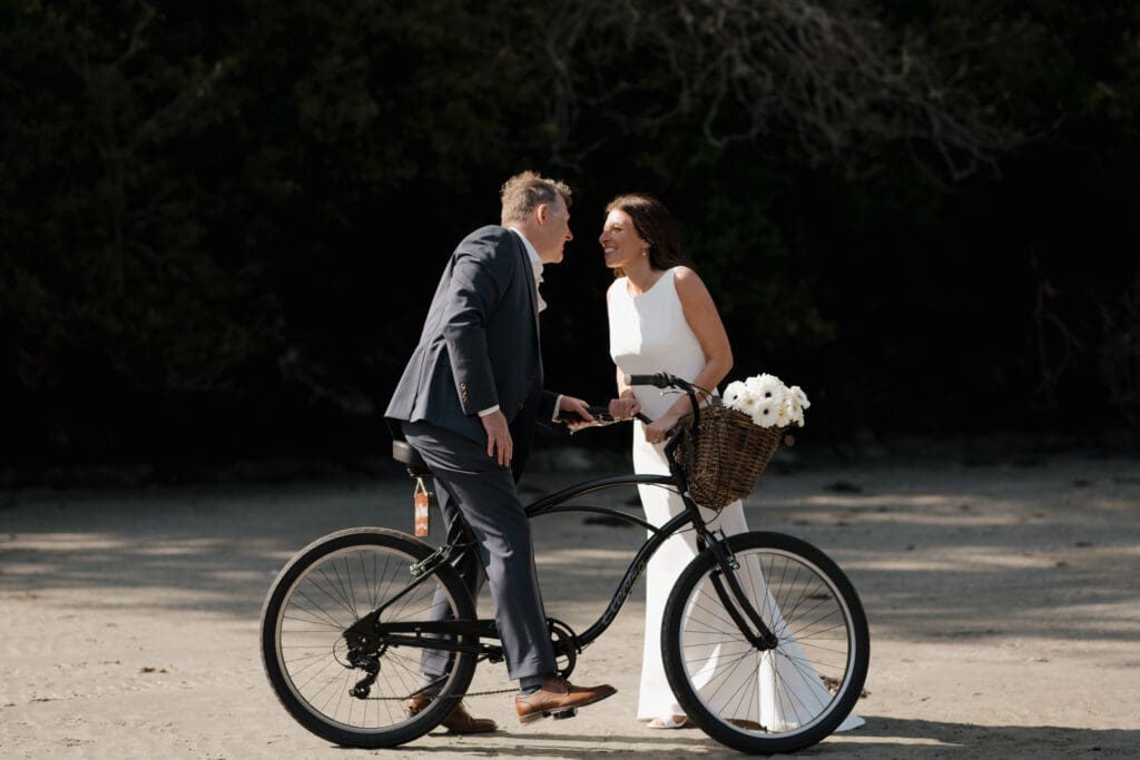 Tofino BC Wedding Photographer and Videographer captures bride and groom after their wedding ceremony at Crystal Cove Resort on Chesterman Beach. They lean in for a kiss overtop a bicycle that they are about to ride down the beach together.