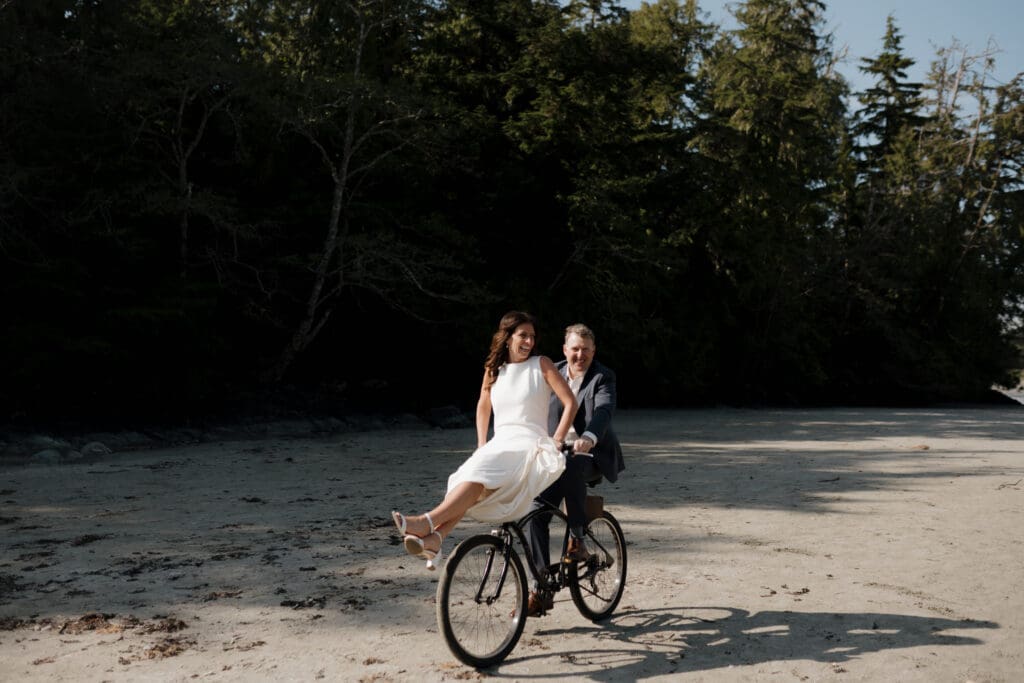 Tofino BC Wedding Photographer and Videographer captures bride and groom after their wedding ceremony at Crystal Cove Resort on Chesterman Beach. They are on a bicycle riding down the beach together.