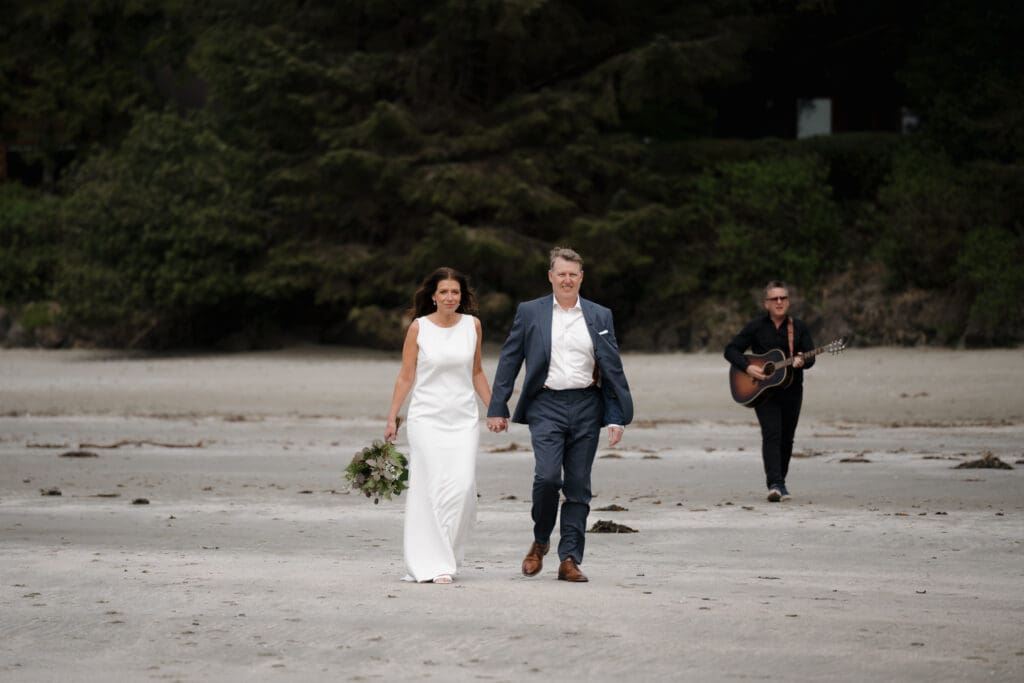 Hayley Zumkeller photography and videography team capture a bride and groom as they walk hand in hand on the beach following their first look, toward the ceremony on Chesterman Beach in Tofino BC on Vancouver Island.