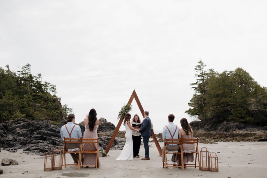 Hayley Zumkeller photography and videography team from Victoria BC capture the intimate elopement beachside ceremony of a bride and groom. We see their A-frame arch behind the officiant and the 4 family member guests looking on.