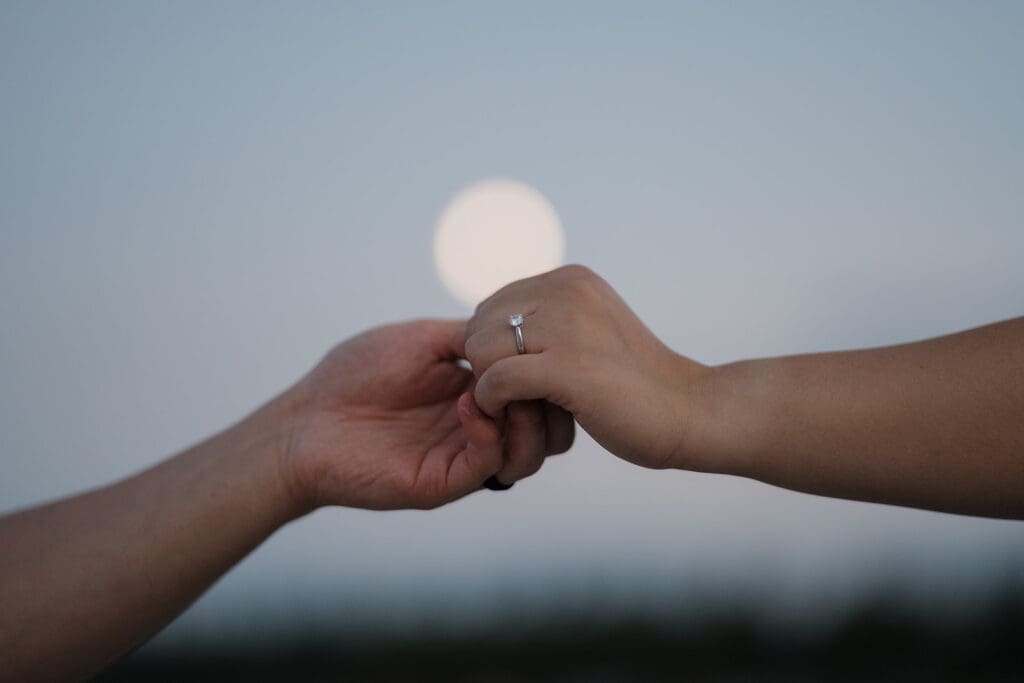Hayley Zumkeller Victoria BC Proposal Photographer captures a couple holding hands with the setting sun in the background, showing off the ring after a private proposal on the beach