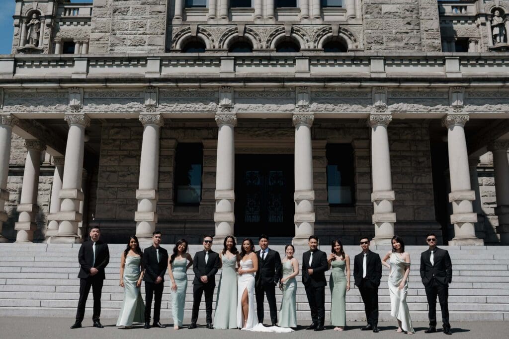 Hayley Zumkeller, Victoria BC Wedding Videographer and Photographer captures a wedding party as they pose outside Parliament on Vancouver Island. The bridesmaids are wearing elegant light green dresses and the groomsman look handsome in their traditional tuxedos.