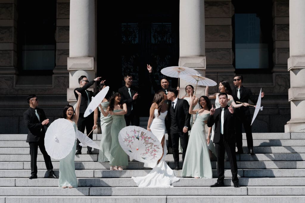 Hayley Zumkeller, Victoria BC Wedding Videographer and Photographer captures a bride and groom kissing surrounded by their wedding party on Parliament grounds in Victoria BC. The bridesmaids all have parasols and are wearing light green gowns.