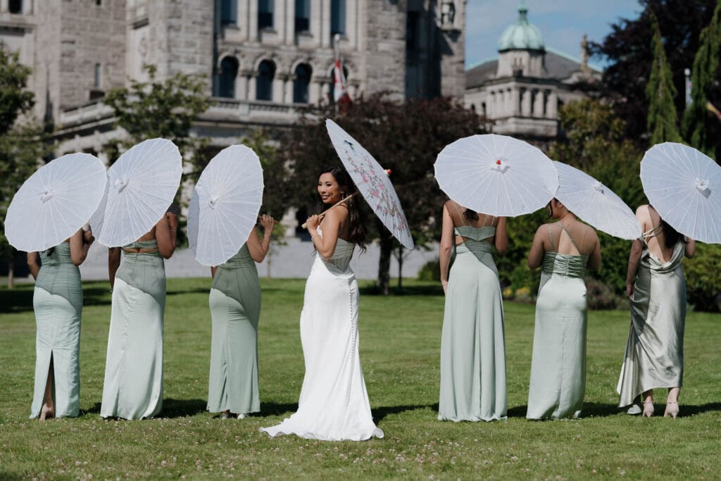 Hayley Zumkeller, Victoria BC Wedding Videography and Photography team capture a bride with her bridesmaids holding parasols on the Parliament Grounds in Victoria BC. The bride is turning back to the camera, but the bridesmaids have their back to the camera with parasols in full view.