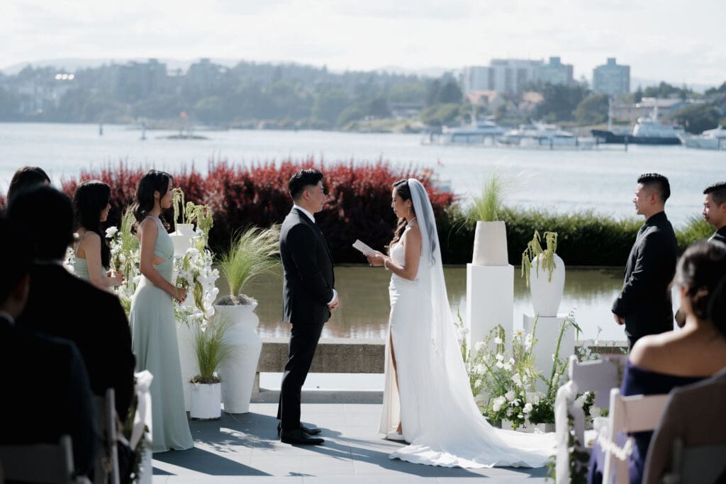 Hayley Zumkeller, Victoria BC Wedding Videographer and Photographer captures the ceremony of an Asian bride and groom as they are reading personal vows at Laurel Point Inn during their mini-destination wedding on Vancouver Island. The harbour view is in the backdrop.