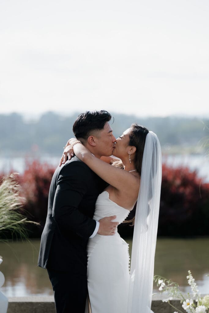 Hayley Zumkeller, Victoria BC Wedding Videographer and Photographer captures the ceremony of an Asian bride and groom as have their first kiss as a married couple at Laurel Point Inn during their mini-destination wedding on Vancouver Island. The harbour view is in the backdrop.