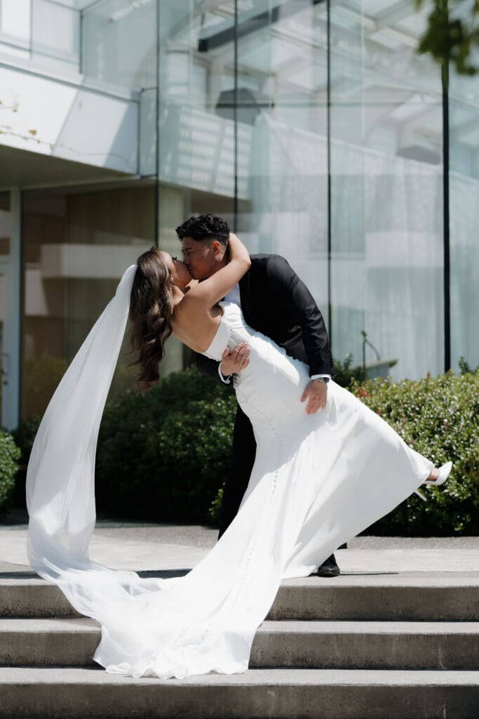 Hayley Zumkeller, Victoria BC Wedding Photographer and Videographer, captures the groom dipkissing the bride after the first look at Laurel Point Inn. The floor to ceiling windows of the reception space are behind them.