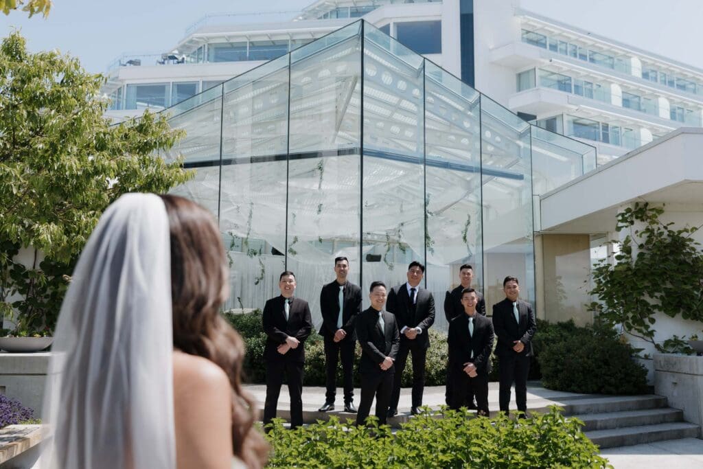 Hayley Zumkeller, Victoria BC Wedding Videographer and Photographer captures a bride in the forefront looking at the groom with his groomsman during wedding party photos at Laurel Point Inn on Vancouver Island.