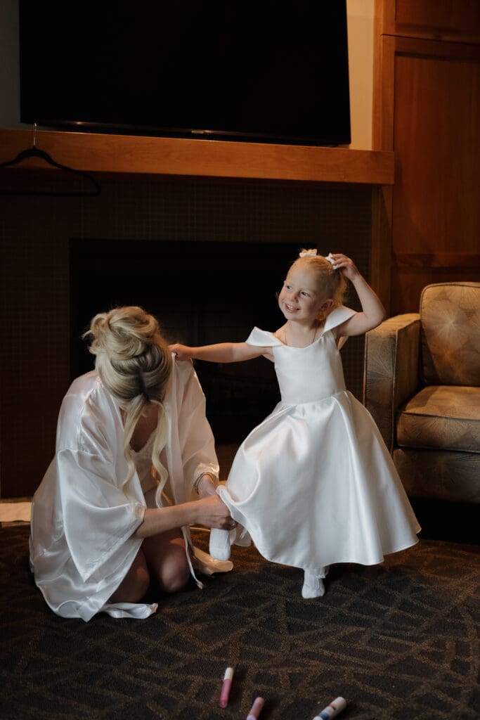 Hayley Zumkeller photography and videography team from Victoria BC capture an image of a bride helping her daughter get her shoes on prior to the ceremony. The bride is still in her white satin robe with hair ready to go. The daughter is dressed in her white satin dress with white bows in her hair.