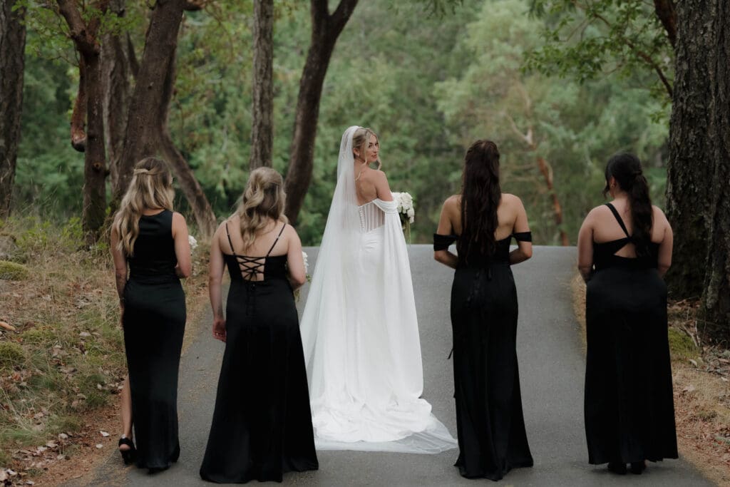 Hayley Zumkeller photography and videography team from Victoria BC capture an elegant image of a bride with her bridesmaids among the tree at Olympic View Golf Course. The bride is turning to look back to the bridesmaids.