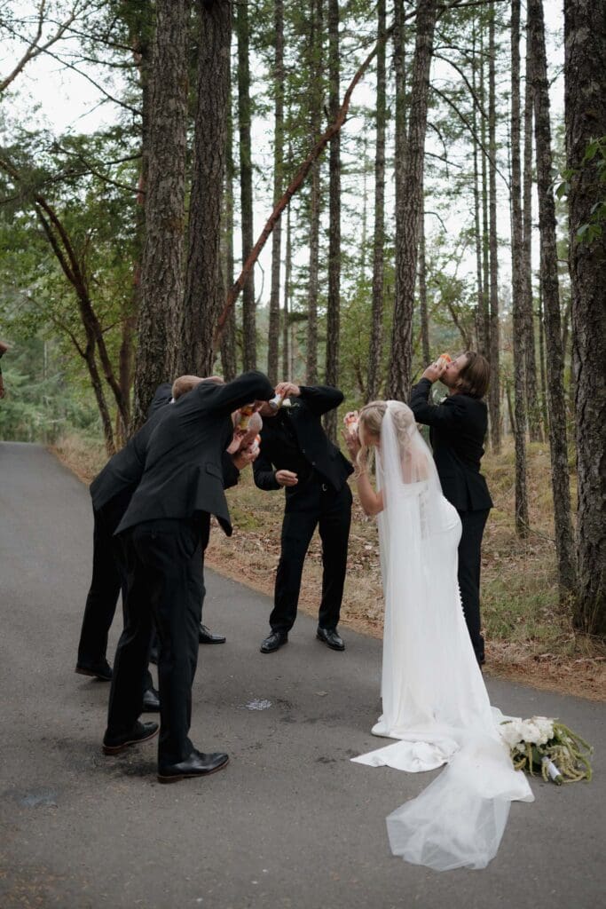 Hayley Zumkeller photography and videography team from Victoria BC capture a candid image of a bride with the groomsman among the trees at Olympic View Golf Course as they all chug a drink.
