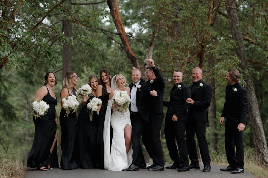 Hayley Zumkeller photography and videography team capture the bride and groom surrounded by the wedding party set among the trees at Olympic View golf course in Victoria BC on Vancouver Island. The bridesmaids dressed elegantly in black gowns and the groomsman in their tuxedos.