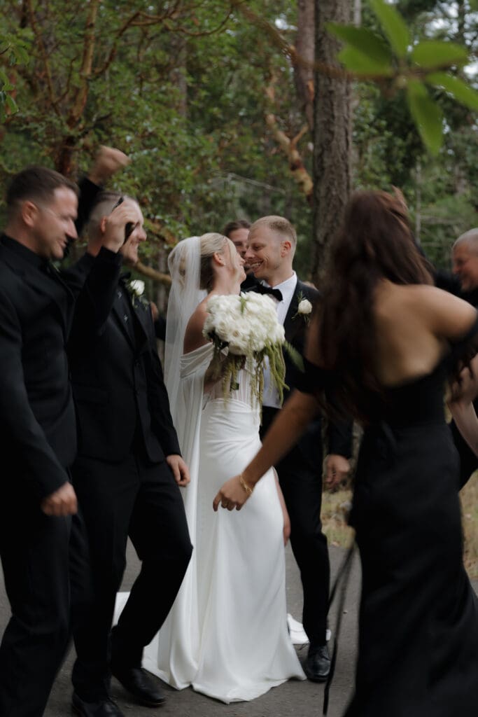 Hayley Zumkeller photography and videography team capture the bride and groom as they lean in for a kiss surrounded by the wedding party cheering them on set among the trees at Olympic View golf course in Victoria BC on Vancouver Island.