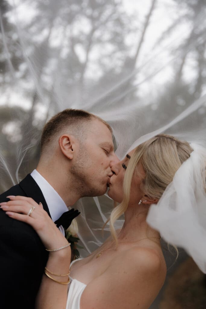 Hayley Zumkeller photography and videography team from Victoria BC capture an elegant image of a bride and groom under the veil kissing among the trees at Olympic View Golf Course.