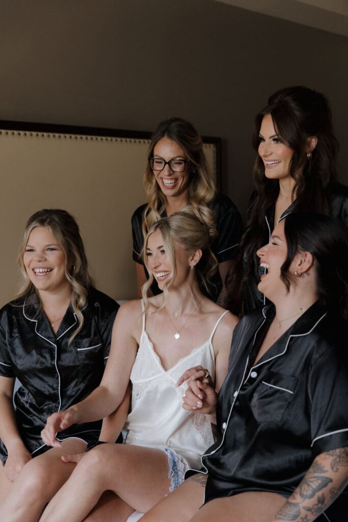 Hayley Zumkeller photography and videography team from Victoria BC capture getting ready photos for a wedding day at Olympic View Golf Course. In this image the bride in white satin pajamas is surrounded by her bridesmaids in their black matching pajamas, all laughing and having a great time together pre-ceremony.
