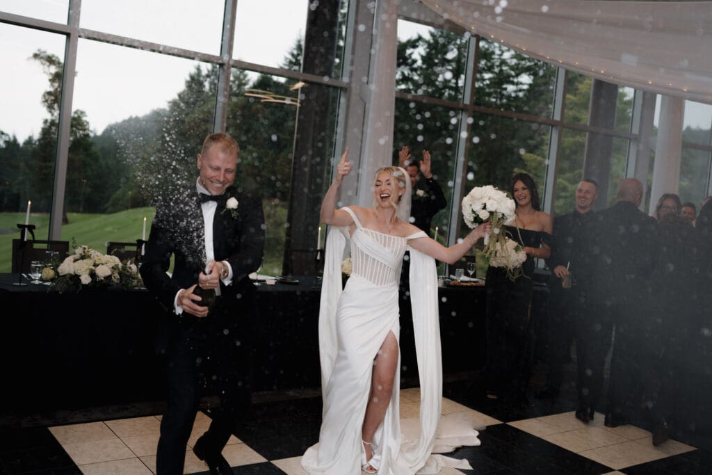 Hayley Zumkeller Photography and Videography team from Victoria BC capture a bride and groom as they arrive to their reception at Olympic View Golf Course on Vancouver Island. The groom is spraying a bottle of champagne while the bride is laughing.