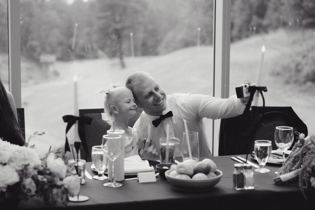 Hayley Zumkeller photography and videography team from Victoria BC capture this black and white image of a groom with his daughter smiling at the head table while taking their photo on the portable cameras. The glass window behind them shows the ground of Olympic View Golf Course on Vancouver Island.