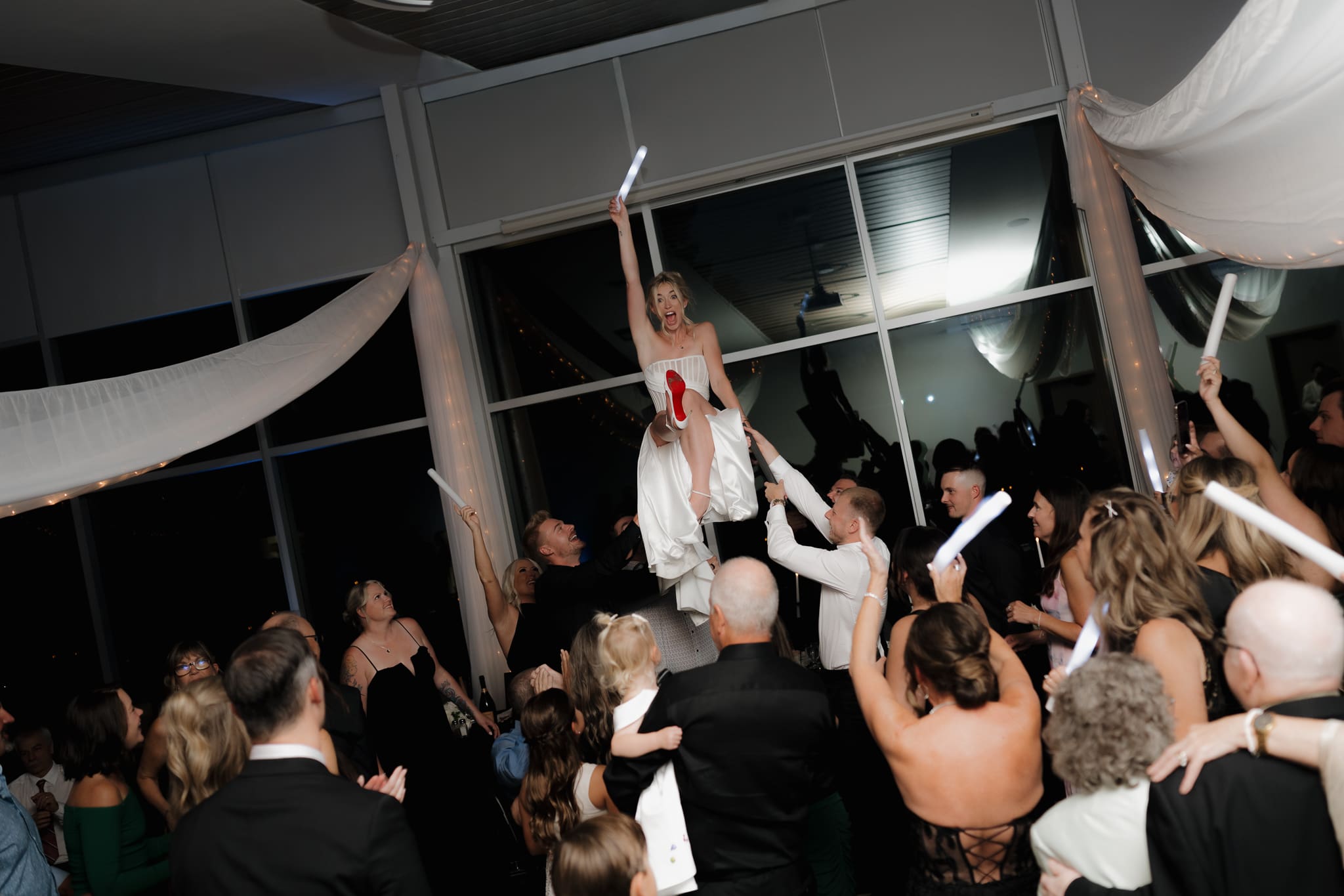 Hayley Zumkeller Photography and Videography team capture a bride laughing and excited, held up in the air on a chair by the groomsman during the reception. The soles of her Louboutin heels flashing the crowd.