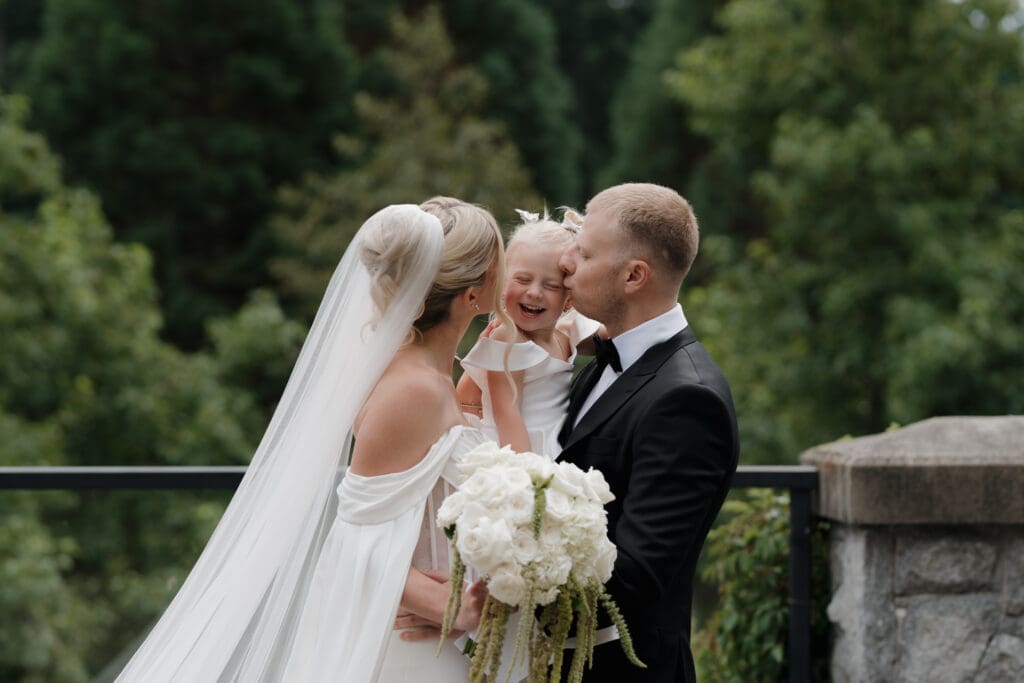 Hayley Zumkeller Photography and Videography team from Victoria BC capture moments just following the first look of a bride and groom with their daughter, each kissing one of her cheeks as she's held up in their mutual embrace. The moment of a family joining together while the greenery of Olympic View Golf Course creates the perfect backdrop. .