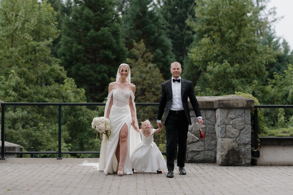 Hayley Zumkeller Photography and Videography team from Victoria BC capture the moment of a family joining together just following the first look of a bride and groom with their daughter, as they each hold one of her hands and look at the camera. The groom holds his daughter's red purse while the greenery of Olympic View Golf Course creates the perfect backdrop.