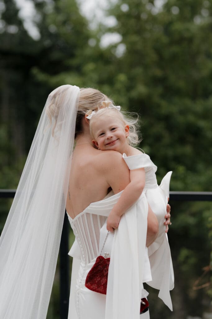 Hayley Zumkeller Photography and Videography team from Victoria BC capture moments just following the first look of a bride and groom with their daughter. The bride is hugging her daughter who is smiling for the camera. The moment of a family joining together.