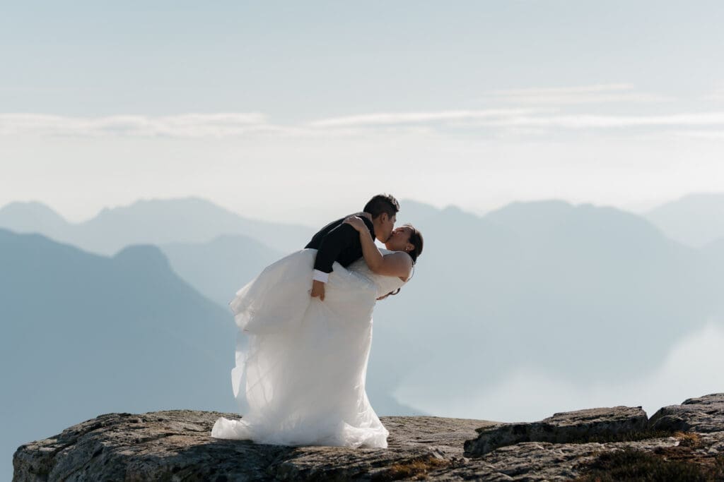 A groom is dip kissing his bride on top of a mountain as part of the images captured by Hayley Zumkeller photography and videography team during this helicopter elopement on Vancouver Island.