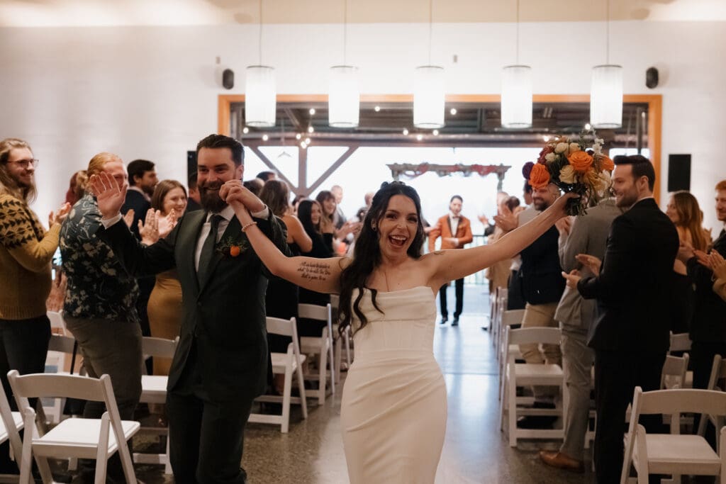 Hayley Zumkeller photography and videography team from Victoria BC capture a newly married couple as they exit the ceremony at Church and State Winery, with celebratory clasped hands in the air. Guests in the background clapping them down the aisle. 