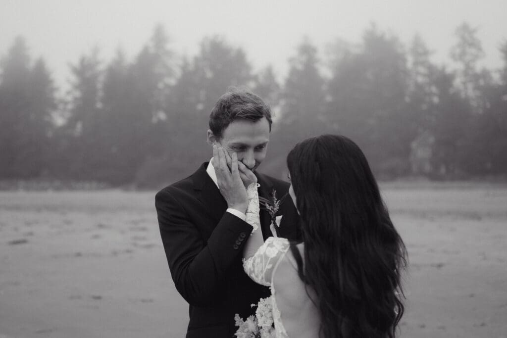 An intimate beachside elopement on Vancouver Island is captured by Hayley Zumkeller photography and videography team. This black and white image from the couple's first look shares an emotional moment where the groom has the bride's hand to his lips as he looks lovingly into her eyes.