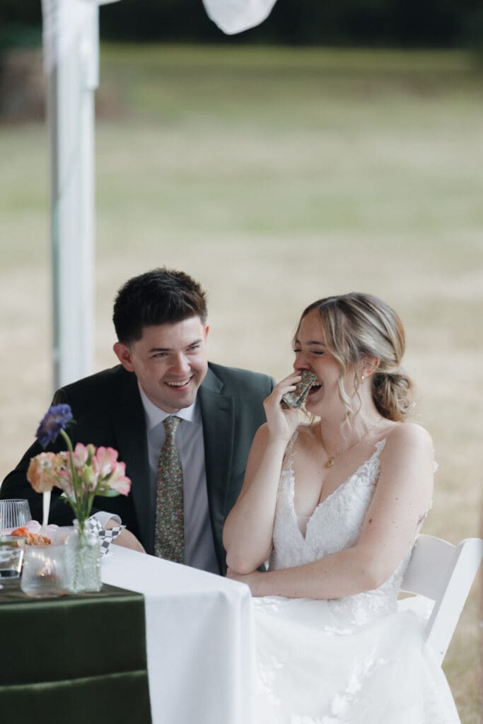 Hayley Zumkeller photography and videography team from Victoria BC capture a newly married bride and groom at their reception listening to the speeches. They are both laughing, the bride has her hand to her face. The lush Pacific Northwest greenery is in the background. 