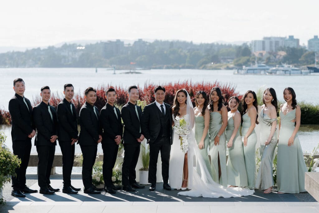 A bride and groom in their elegant wedding attire pose for photos with their wedding party. On Vancouver Island there are many vendors that specialize in wedding gowns and suits to cater to all styles. Image captured by Hayley Zumkeller photography and videography team.