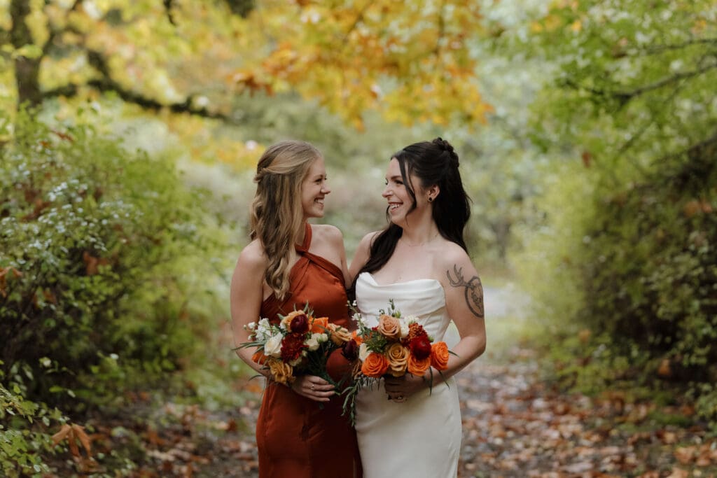 Hayley Zumkeller photography and videography team from Victoria BC captures a bride looking at one of her bridesmaids - dressed in vibrant orange for this fall wedding - in the forest at Church and State Winery on Vancouver Island.