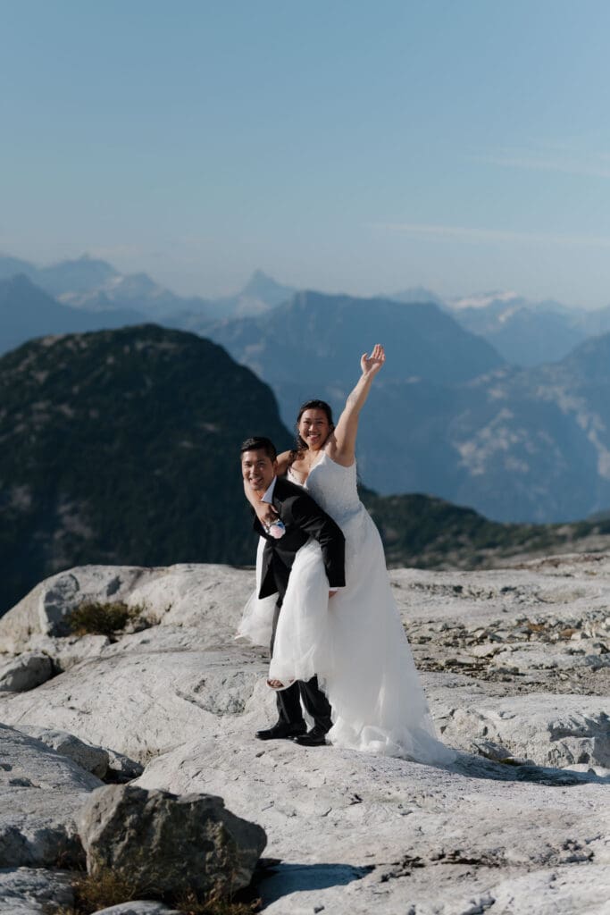 Romantic close-up of couple laughing and having fun during their mountaintop helicopter elopement on Vancouver Island. In the image, the bride is piggy back on her husband waving at the camera with the mountain views in the backdrop.