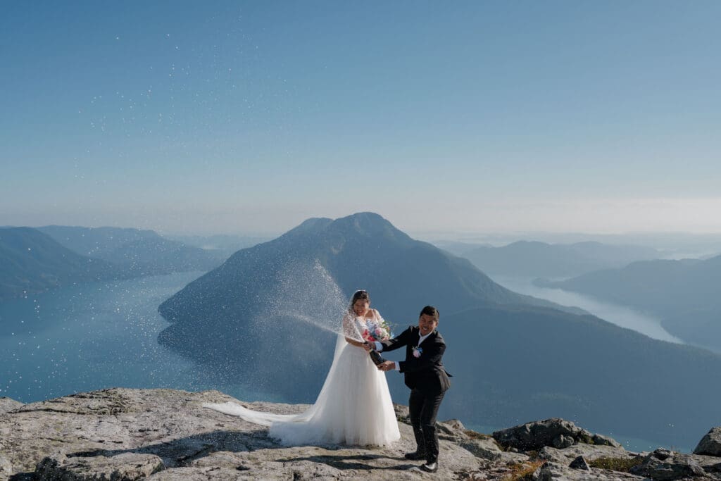 A champagne bottle sprays as it's being opened by a newly married bride and groom during their mountaintop elopement on Vancouver Island with cinematic mountain and water views in the background.