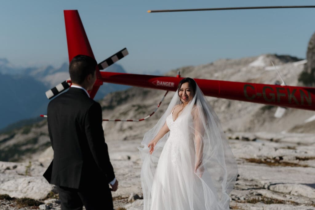 Bride displaying her dress for her groom on a mountain cliff during their Vancouver Island helicopter elopement with scenic views. You can see the back of the red helicopter in the background.