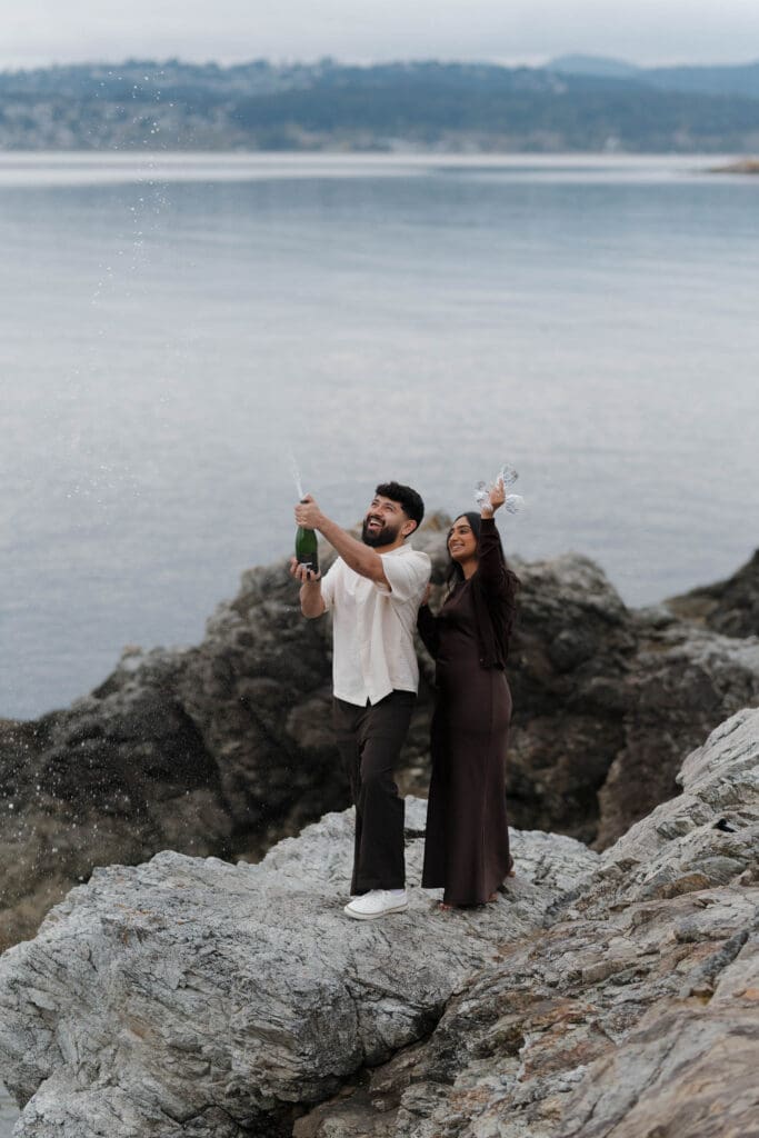 Newly engaged couple celebrating with champagne at Saxe Point Park, Victoria BC — proposal and engagement photography by Hayley Zumkeller