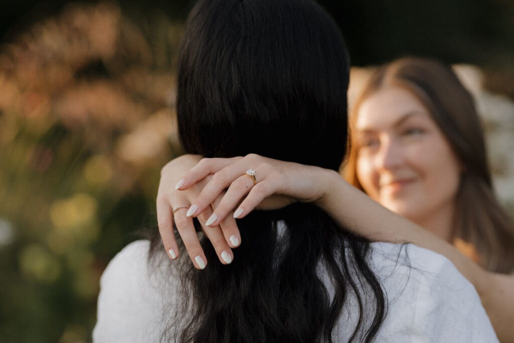 The engagement ring is displayed on the hand of a newly engaged couple as it clasps her partner around the neck in an intimate pose.
