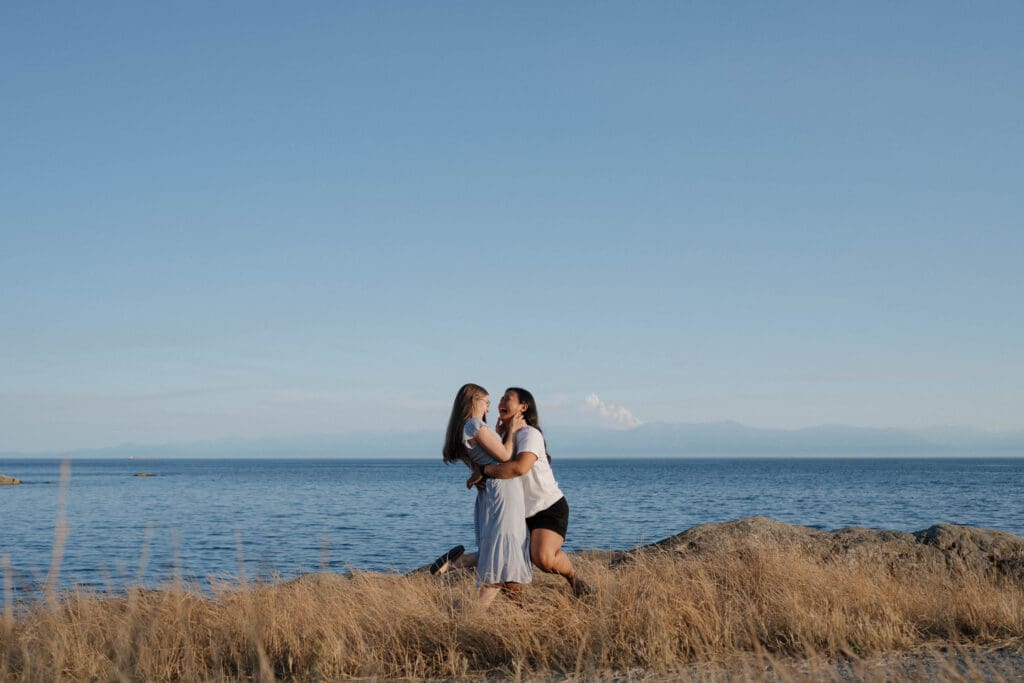 Couple running together on the oceanfront path at Saxe Point Park after a surprise proposal session in Victoria BC during golden hour on Vancouver Island