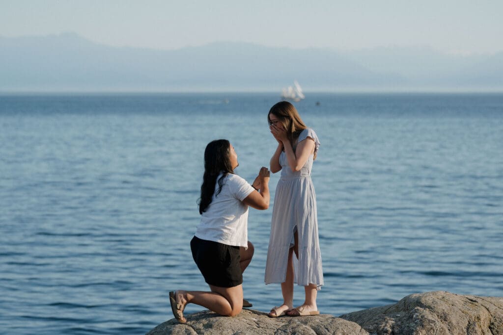 Surprise proposal at Saxe Point Park in Victoria BC on the rocks by the ocean at sunset with sailboat and mountains in the background
