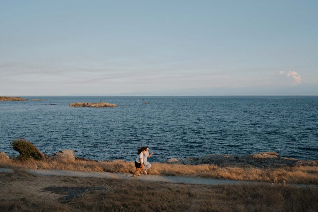 Couple running together on the oceanfront path at Saxe Point Park after a surprise proposal session in Victoria BC during golden hour on Vancouver Island