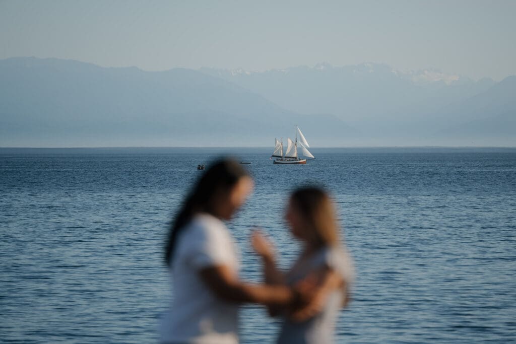 Creative blurred portrait of engaged couple on rocky shoreline at Saxe Point Park with sailboat in focus during a proposal session in Victoria BC