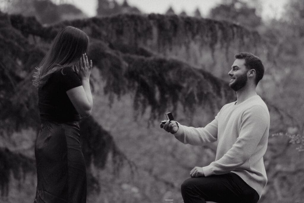 Black and white image of a man on one knee proposing to partner in the public gardens of Government House Victoria, captured by hidden photographer.