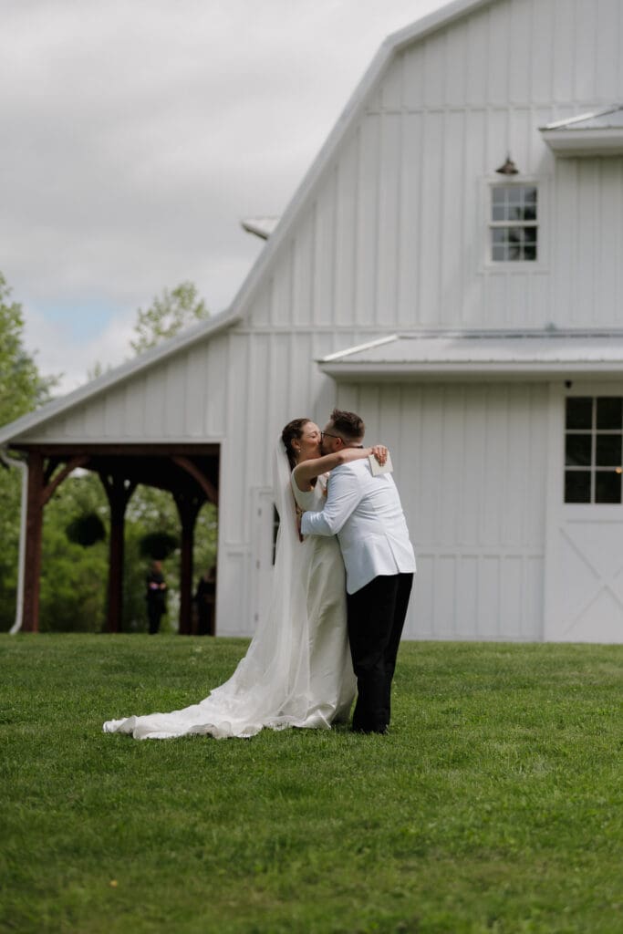 A bride and groom during their first look locked in an embrace. In the background is the white barn found on the estate of Wildflower on Watts in Honesdale PA.