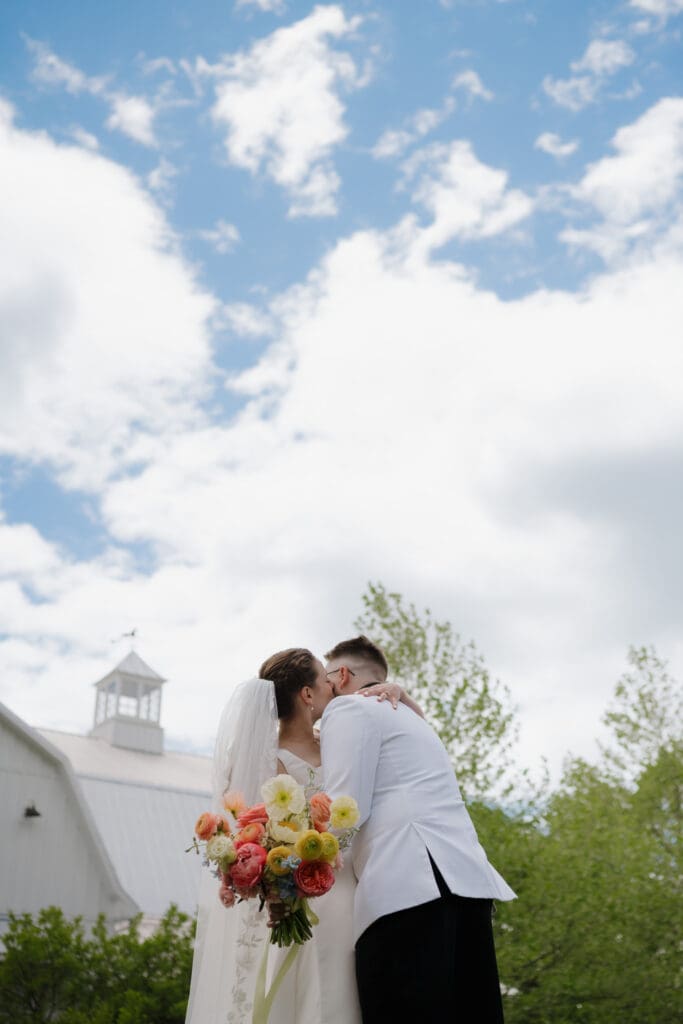 The beautiful blue skies with fluffy white clouds are seen behind a kissing bride and groom during their first look. The top of the white barn at Wildflower on Watts can be seen in the background and the bride holds her colourful floral bouquet.